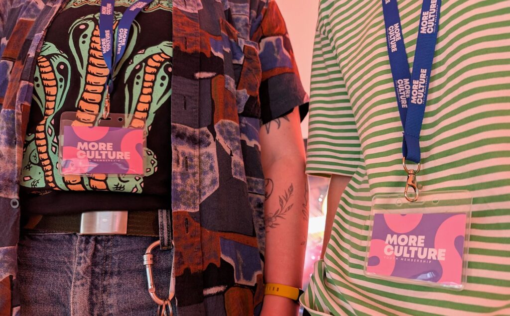Two people wearing colourful outfits with "More Culture" badges. One has a patterned shirt and the other a green striped shirt. Visible tattoos and accessories.
