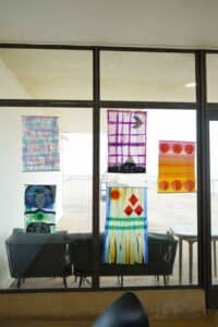 Colourful abstract paintings hang on a glass wall, featuring geometric and linear patterns. A patio chair and table are visible outside, evoking a calm and artistic atmosphere.