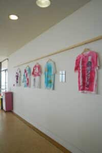 Five vibrantly coloured T-shirts, with tie-dye and graphic designs, hang on a wall in a bright room. The display exudes a creative, artistic vibe.