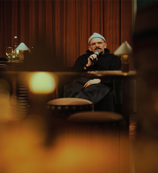 A person wearing a light blue beanie and glasses sits in an armchair, holding a microphone and speaking.